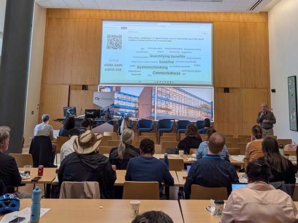 A view inside the Scholars Event Theater of a session of the Sustainability Showcase. A man speaks to a crowd while presenting slides on a large projection screen.