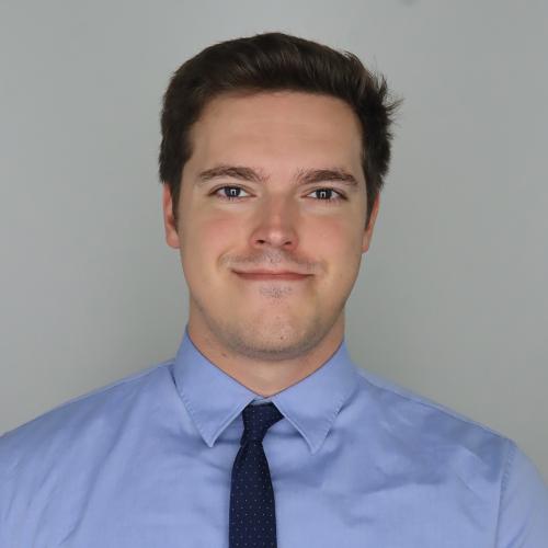 Headshot portrait of an individual photographed from the chest up against a plain light gray background. The individual is wearing a light blue button‑down shirt and a dark patterned tie, facing the camera in a centered studio-style composition. The image is evenly lit with a neutral backdrop and no visible text or additional objects.