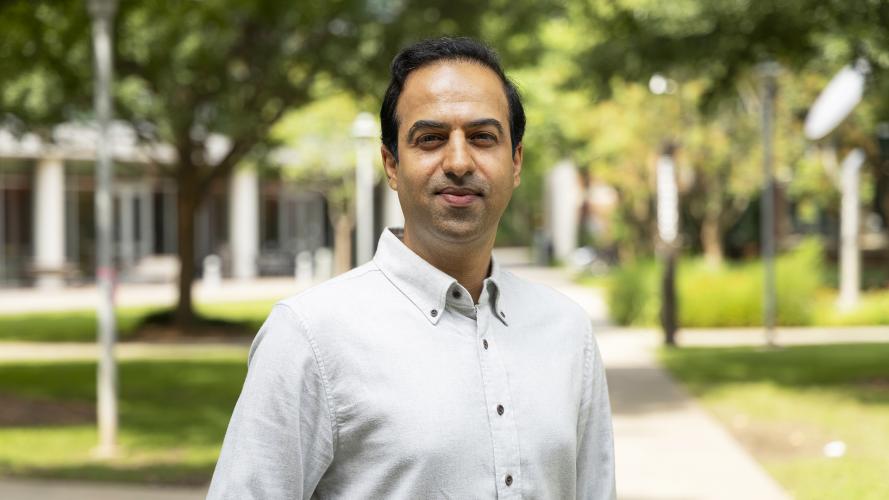 Portrait of an individual standing on a paved campus walkway, wearing a light-colored button-down shirt. Trees, landscaped green spaces, and campus buildings appear in the softly blurred background, with daylight illuminating the outdoor scene. The image is framed from the waist up, with the individual centered in the foreground.