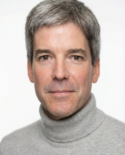 Headshot portrait of an individual photographed from the shoulders up against a plain light gray background, wearing a light gray turtleneck sweater. The individual faces forward with short, neatly styled hair and evenly lit features, centered in the frame.
