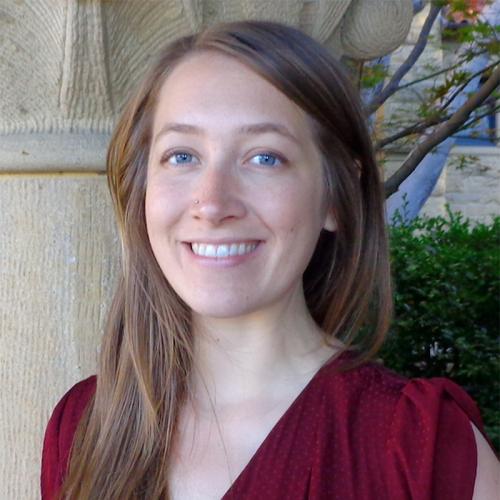 Portrait of an individual photographed outdoors, shown from the shoulders up and wearing a dark red top. The background includes a textured stone column, greenery, and part of a building with a window visible behind the subject.